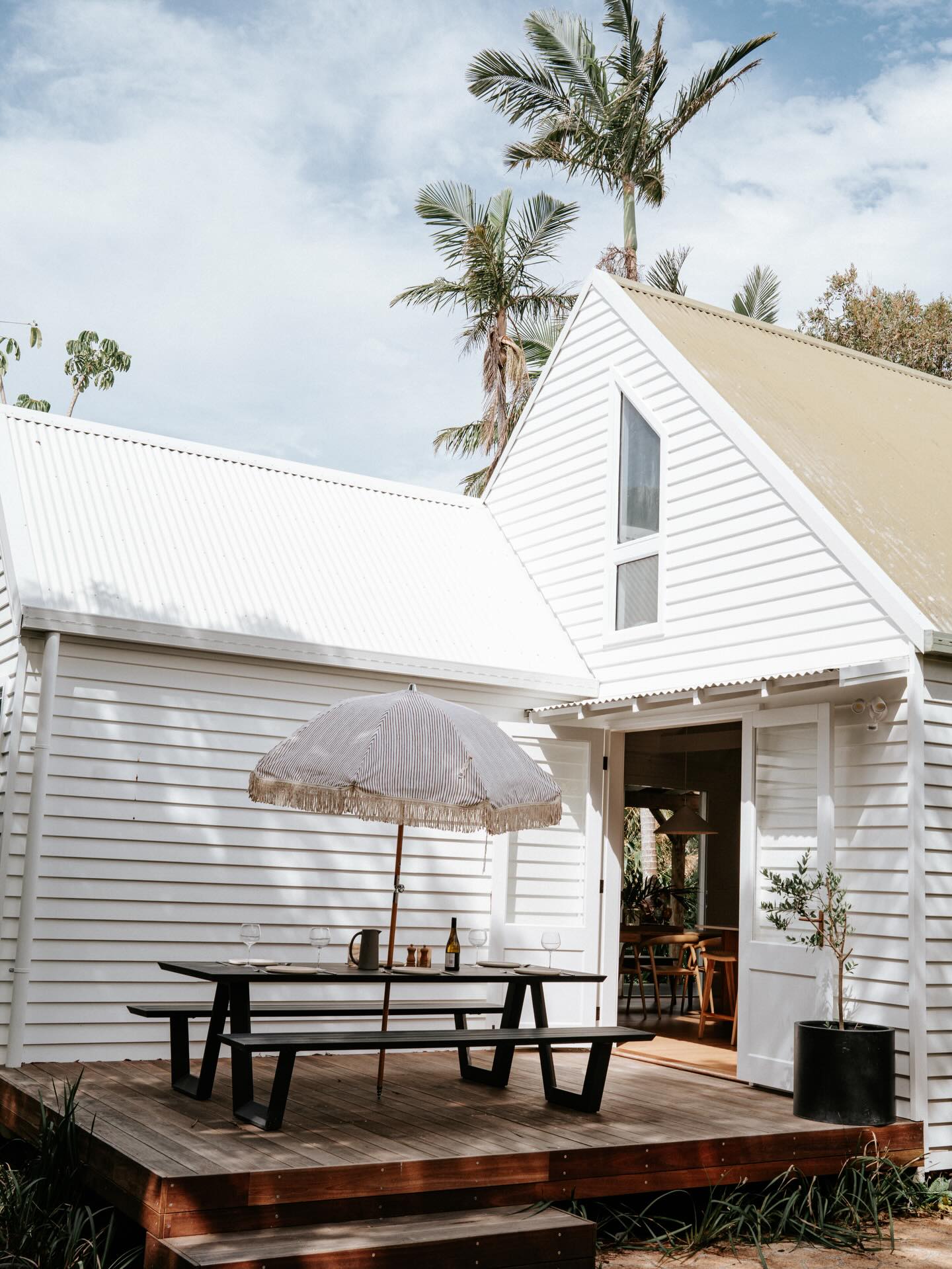 White gabled cottage with a timber deck, picnic table and a canvas umbrella under palms
